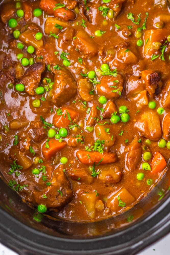 Close-up of beef stew with chunks of beef, carrots, potatoes, and green peas in a thick brown sauce, garnished with chopped herbs.