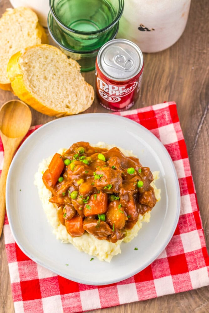A plate of mashed potatoes topped with beef stew, carrots, and peas sits on a red plaid napkin next to a wooden spoon, sliced bread, a glass, and a can of Dr. Pepper.