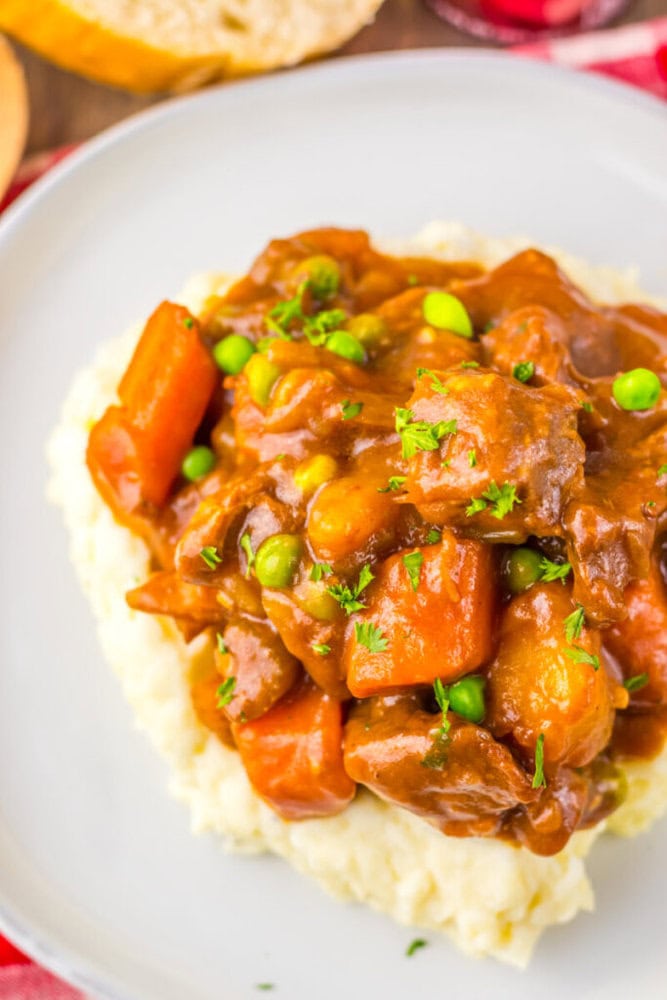A serving of beef stew with carrots, peas, and potatoes in brown gravy is placed on top of mashed potatoes on a white plate.