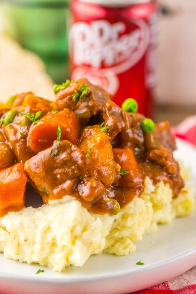 Beef stew with carrots, peas, and gravy served over mashed potatoes on a white plate, with a can of Dr Pepper in the background.