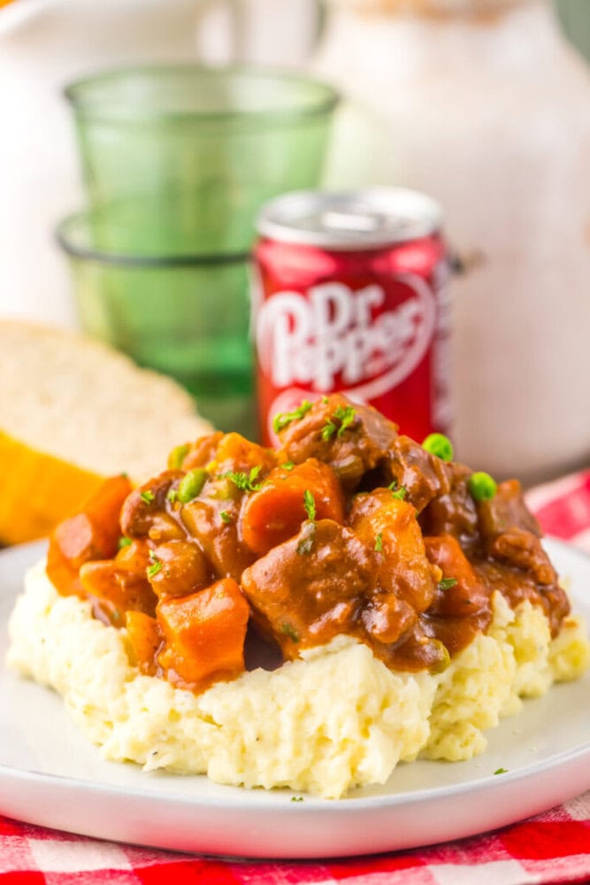 A plate of mashed potatoes topped with beef stew and vegetables, with a can of Dr Pepper and bread in the background.