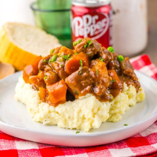 A plate of mashed potatoes topped with beef stew, garnished with parsley, beside sliced bread and a can of Dr Pepper on a red checkered cloth.