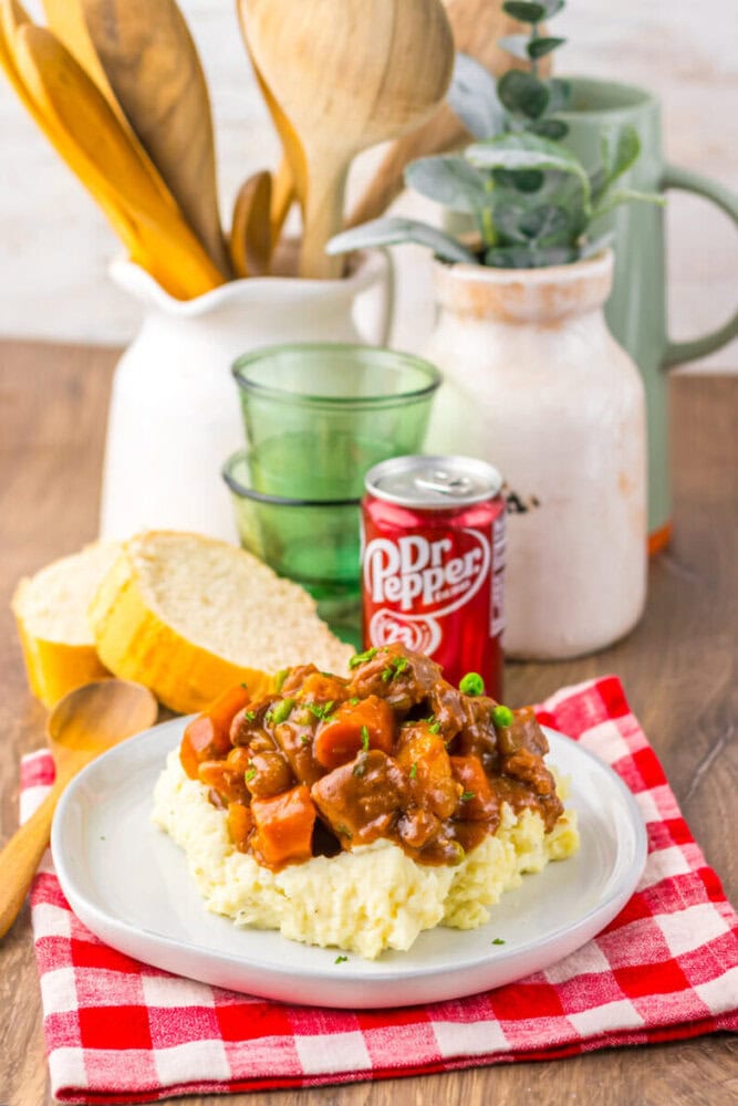A plate of mashed potatoes topped with stew sits on a red checkered cloth, next to sliced bread, a can of Dr Pepper, glass cups, and kitchenware in the background.