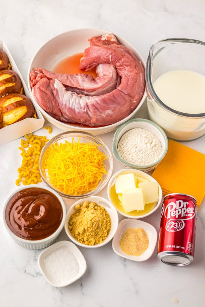 Various ingredients are arranged on a counter, including raw pork tenderloin, shredded cheese, milk, flour, butter, barbecue sauce, macaroni, Dr Pepper, pretzel buns, and seasonings.