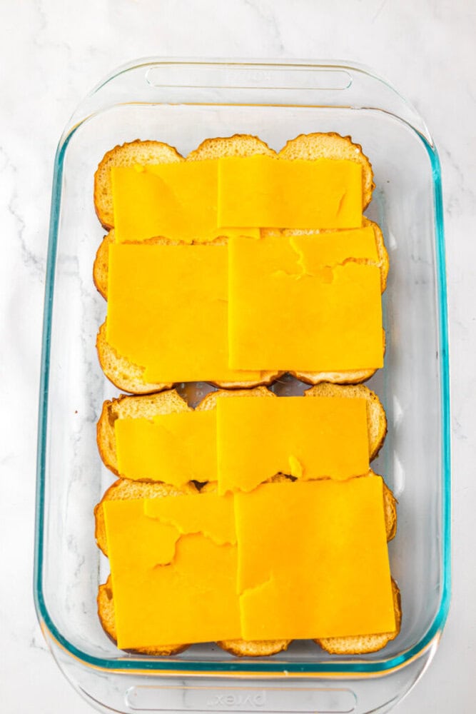 Slices of cheddar cheese laid on top of halved sandwich rolls arranged in a glass baking dish on a white surface.