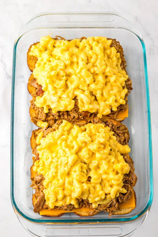 A glass baking dish holding two open-faced sandwiches topped with pulled meat, cheddar cheese slices, and macaroni and cheese.