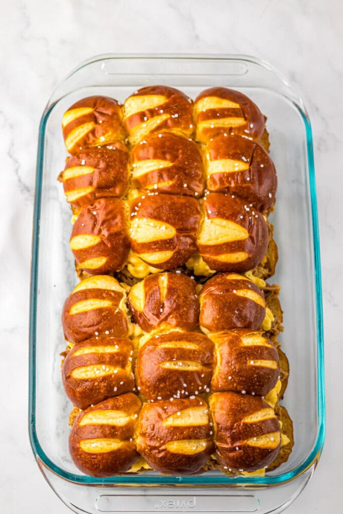 A glass baking dish filled with neatly arranged, golden-brown pretzel buns topped with coarse salt and filled with melted cheese.