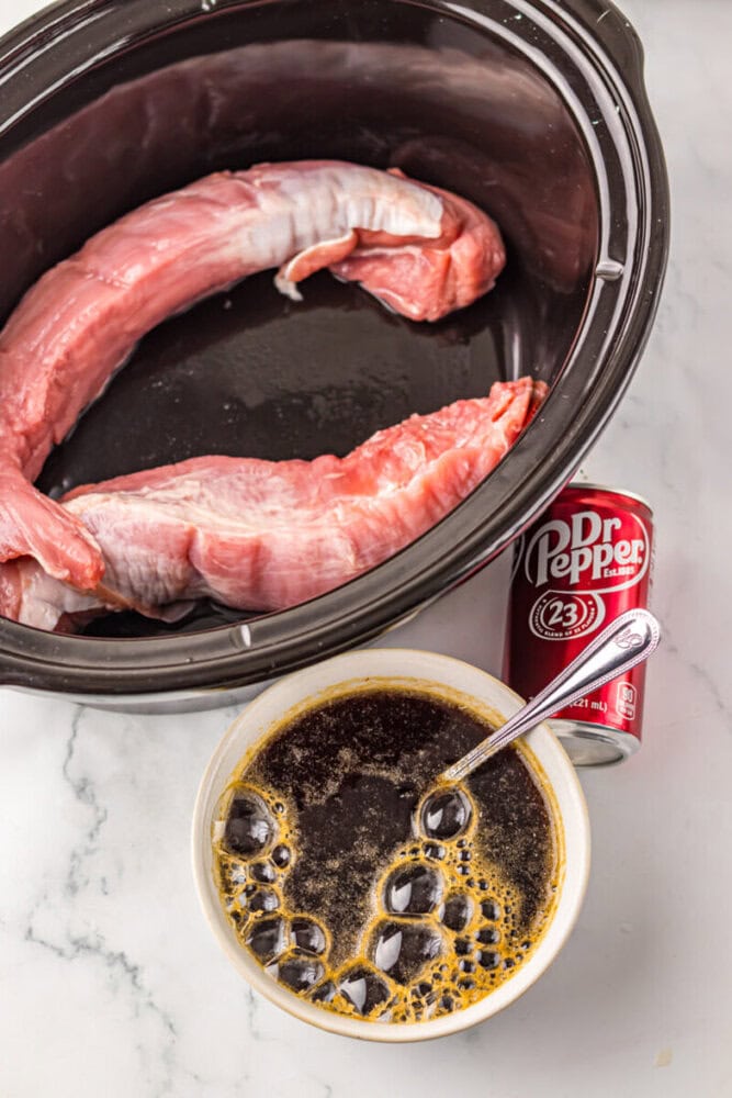 Two raw pork tenderloins in a slow cooker, with a can of Dr Pepper and a bowl of dark marinade with a spoon on a white surface.