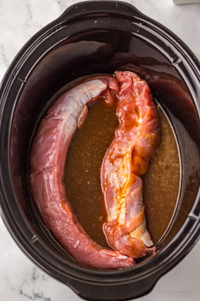 Two raw pork tenderloins sit in marinade inside a black slow cooker on a white countertop.