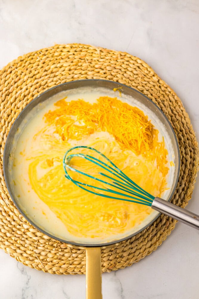 A pan with milk and shredded cheddar cheese being whisked on a woven placemat, ready to make a cheese sauce.