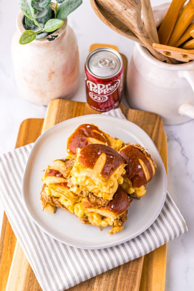 A plate with three pretzel bun sandwiches filled with shredded meat and macaroni and cheese, next to a can of Dr Pepper and kitchen utensils on a striped cloth.