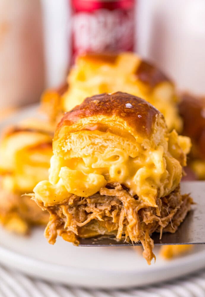 A close-up of a slider sandwich filled with pulled pork and macaroni and cheese on a pretzel bun, held up on a spatula.