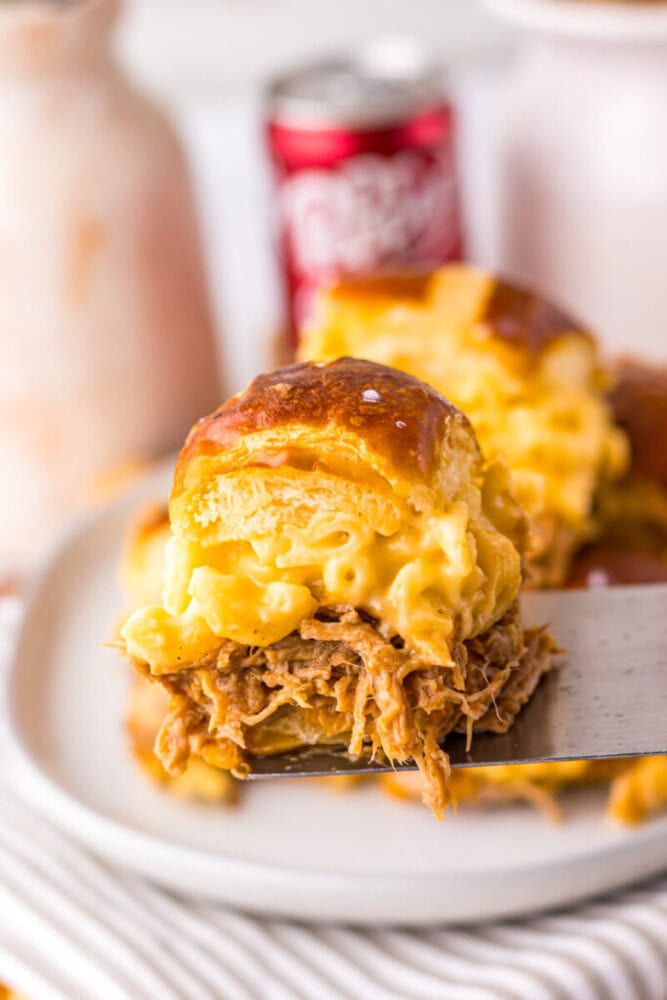 A close-up of a sandwich with pulled pork and macaroni and cheese on a pretzel bun, being served on a spatula with a can of Dr Pepper in the background.