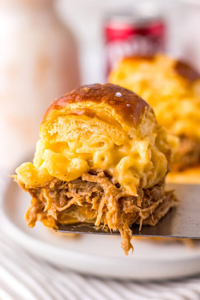 A close-up of a slider sandwich filled with pulled pork and macaroni and cheese on a pretzel bun, held on a spatula.
