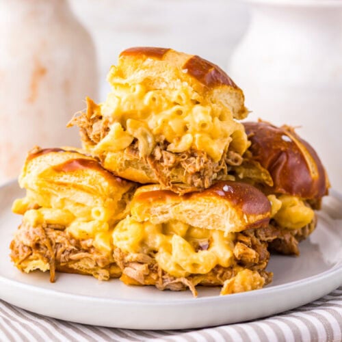 Three pulled pork and macaroni and cheese sliders on pretzel buns are stacked on a plate with a striped napkin, with white pitchers in the background.