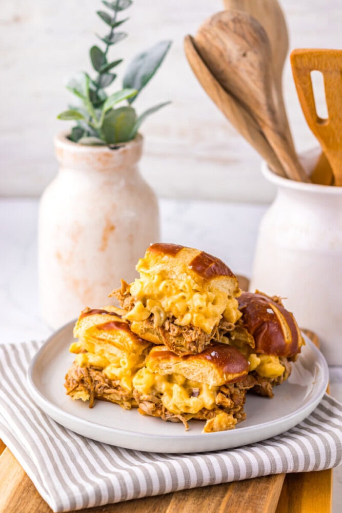 A plate of pulled pork and macaroni and cheese sliders on pretzel buns, stacked on a striped napkin with ceramic jars and wooden utensils in the background.