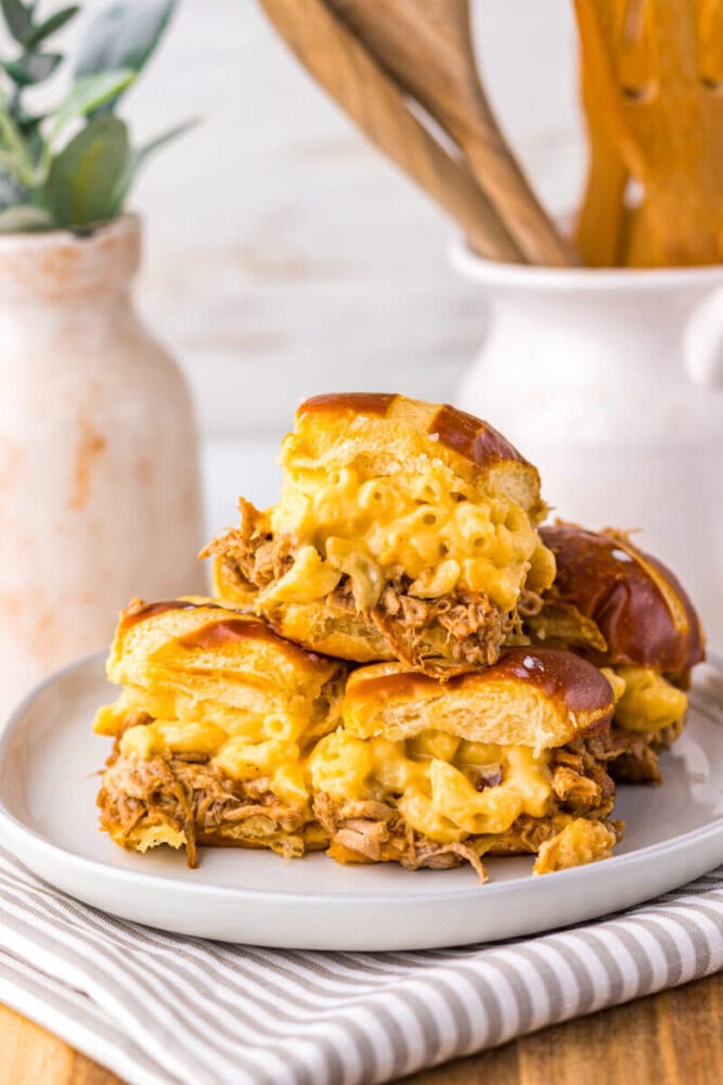 Three slider sandwiches filled with pulled pork and macaroni and cheese are stacked on a white plate, with a striped napkin underneath and kitchen decor in the background.