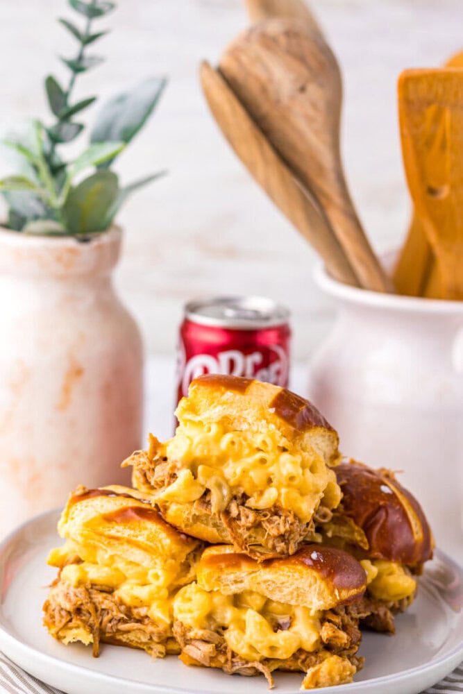 A stack of sandwiches filled with pulled pork and macaroni and cheese on a plate, with a can of Dr. Pepper and kitchen utensils in the background.