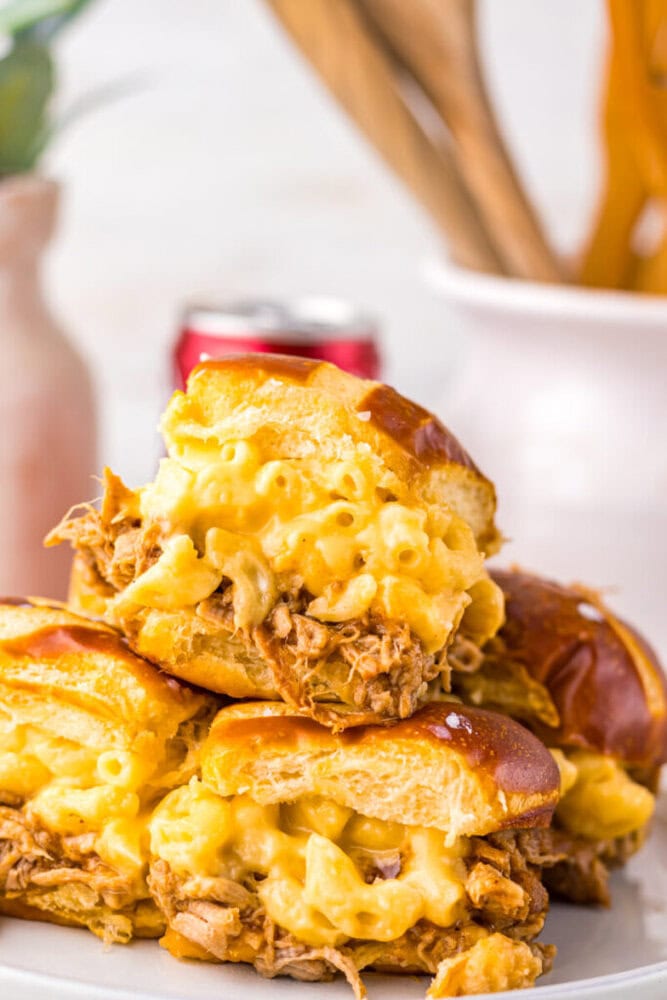 Three stacked sliders filled with shredded meat and macaroni and cheese on a white plate, with blurred kitchen utensils in the background.
