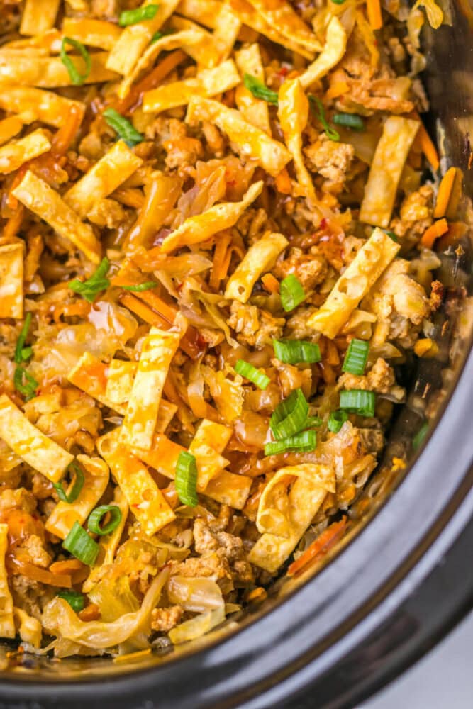 A close-up of an egg roll in a bowl dish with ground meat, sliced cabbage, carrot strips, fried wonton strips, and chopped green onions inside a slow cooker.