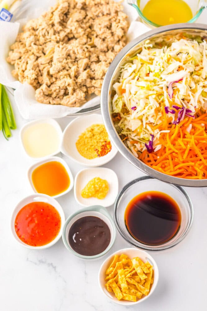 Overhead view of a bowl with shredded cabbage and carrots, surrounded by small bowls of sauces, seasonings, cooked meat, and crispy strips on a white surface.