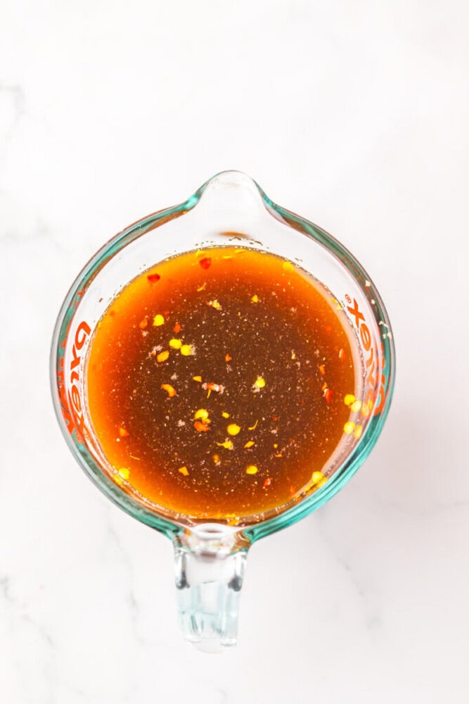A glass measuring cup filled with a reddish-brown liquid containing visible chili flakes and spices, placed on a white marble surface.