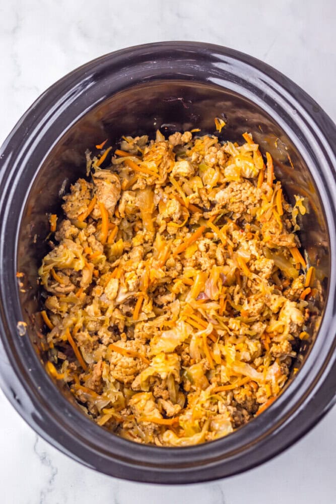 A slow cooker filled with a cooked mixture of shredded cabbage, ground meat, and carrots on a light marble surface.