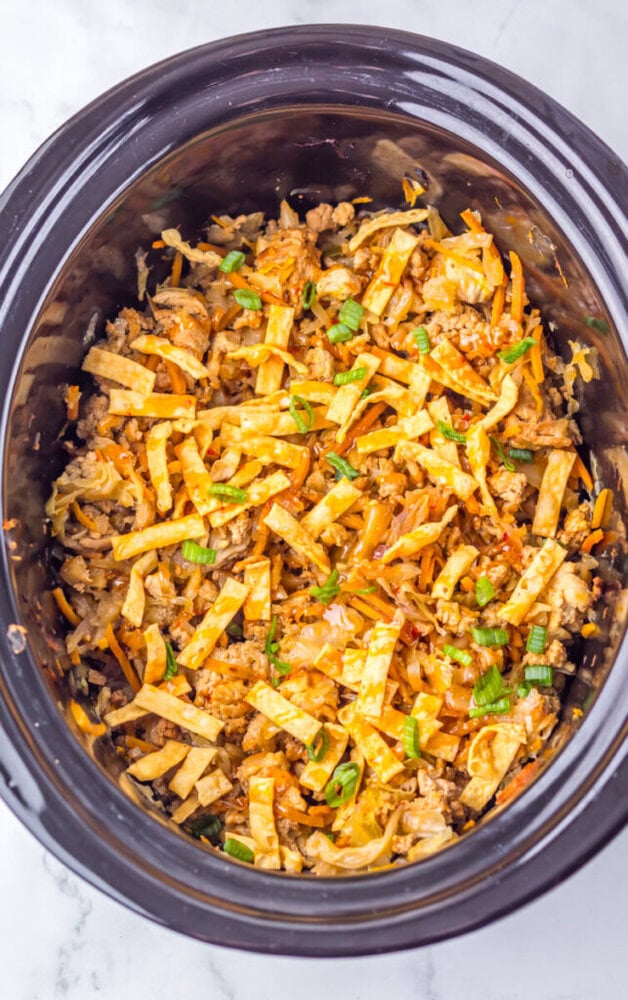 Cooked egg roll in a bowl dish with shredded vegetables, ground meat, and sliced egg strips, garnished with chopped green onions, inside a slow cooker.