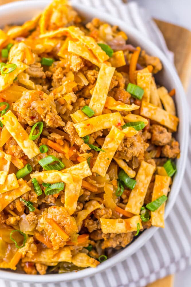 A bowl filled with egg roll in a bowl, featuring ground meat, shredded carrots, cabbage, crispy wonton strips, and chopped green onions on top.
