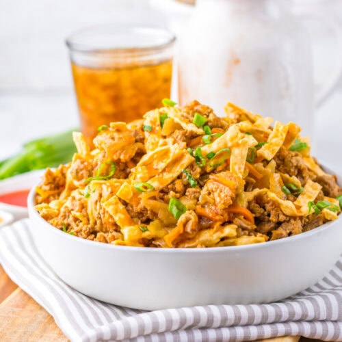 A white bowl filled with stir-fried ground meat, shredded cabbage, and green onions sits on a striped napkin with iced tea and condiments in the background.