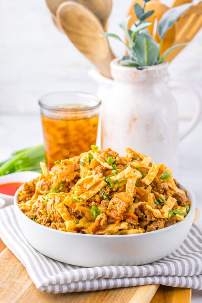 A bowl of taco casserole topped with chopped green onions sits on a striped cloth, with a glass of iced tea and kitchen utensils in the background.