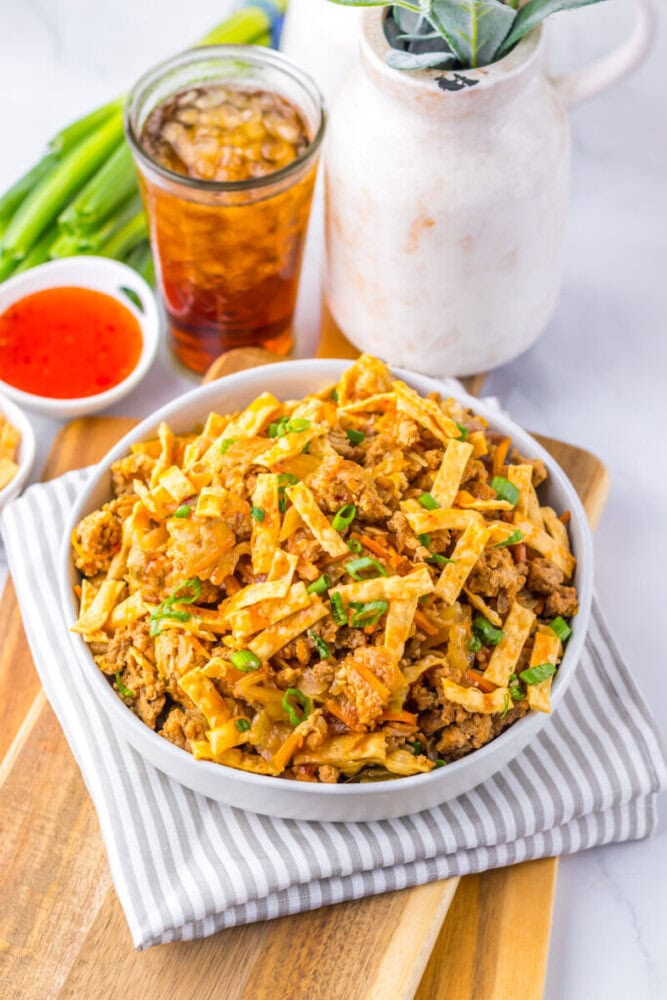 A bowl of egg roll in a bowl with ground meat, cabbage, and crispy wonton strips, garnished with green onions, next to a drink and dipping sauce on a striped napkin.