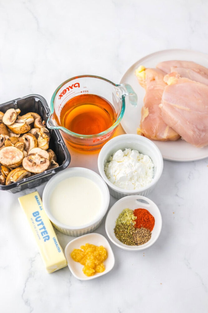 Raw chicken breasts on a plate, sliced mushrooms, butter, chicken broth, cream, garlic, cornstarch, and various spices arranged on a white surface.