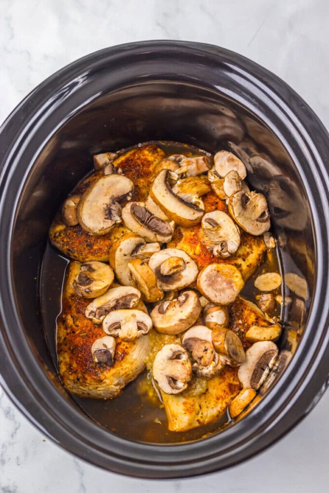 Seasoned chicken breasts and sliced mushrooms in a slow cooker with a small amount of liquid.