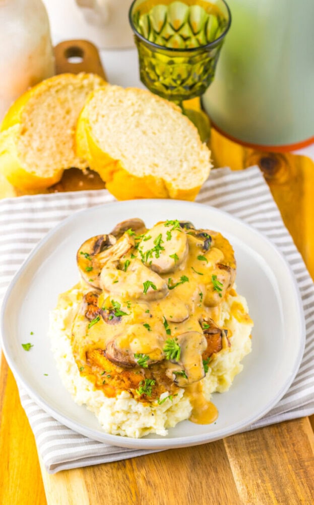 A plate of mashed potatoes topped with chicken and creamy mushroom sauce, garnished with herbs, with sliced bread and a green glass in the background.
