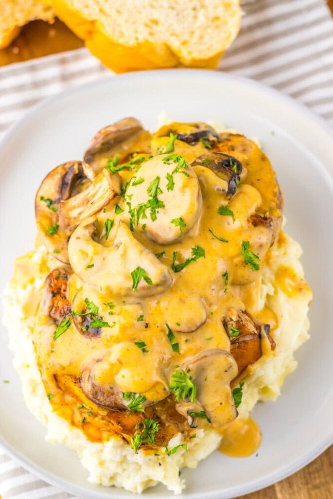 Plate of mashed potatoes topped with a chicken breast, creamy mushroom sauce, sliced mushrooms, and chopped parsley, with a slice of bread in the background.