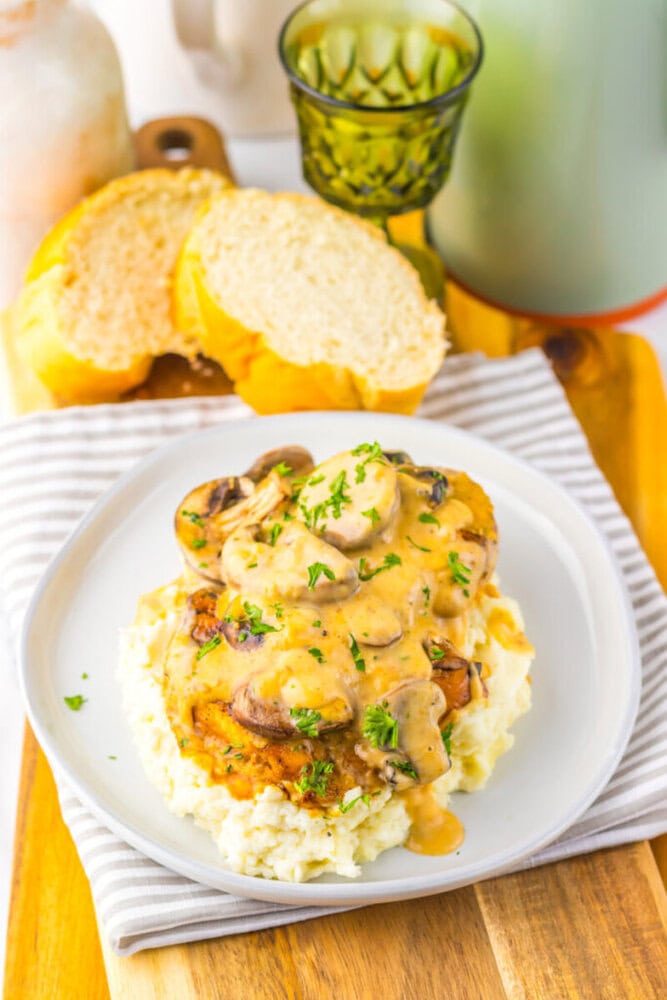 A plate of mashed potatoes topped with chicken, mushrooms, and creamy sauce, garnished with parsley. Slices of bread and a glass of drink are in the background.