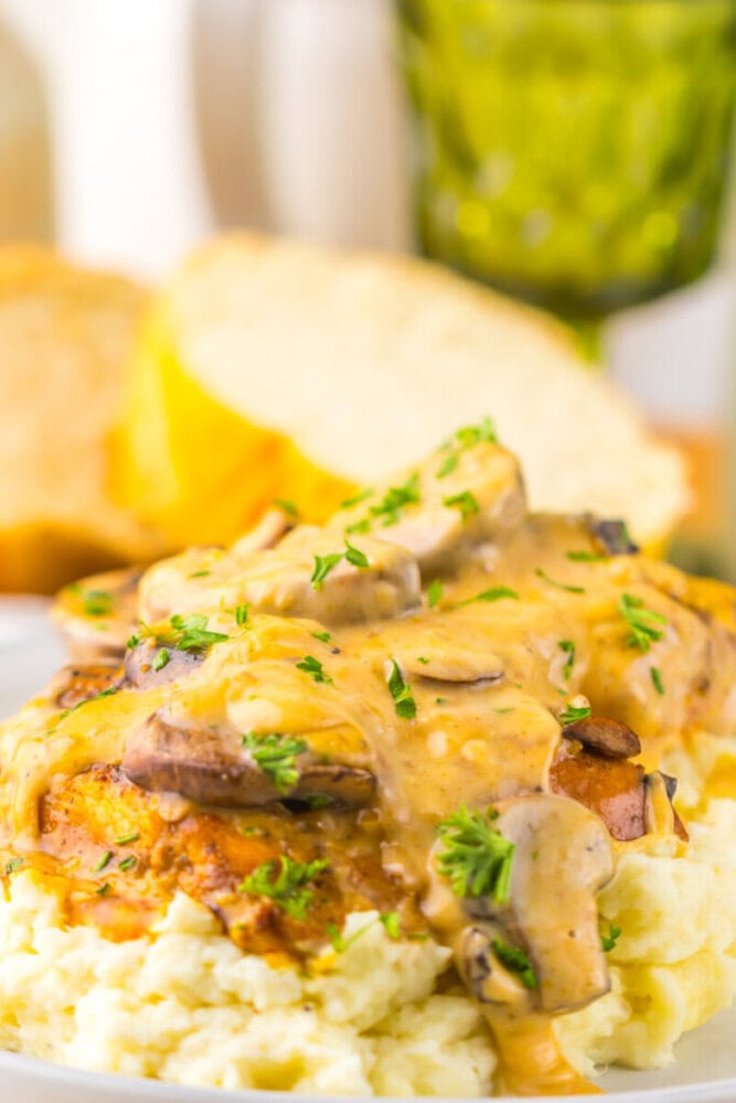 A plate of mashed potatoes topped with a grilled chicken breast, mushroom gravy, and chopped herbs, with bread and a green glass in the background.