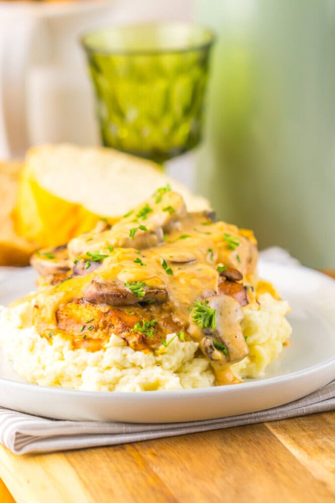 A plate of mashed potatoes topped with grilled chicken breast, mushroom gravy, and fresh herbs, with a slice of bread and a green glass in the background.