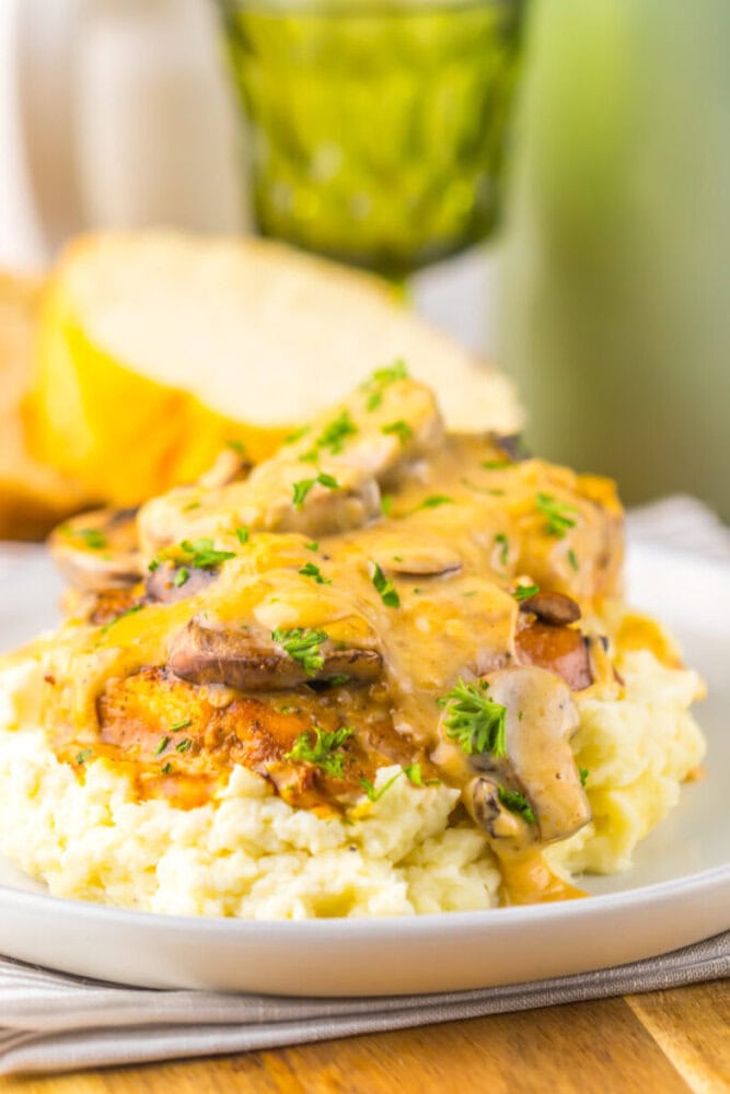 Grilled chicken breast topped with creamy mushroom sauce and chopped herbs, served on a bed of mashed potatoes, with a slice of bread in the background.