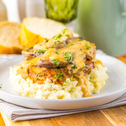 A plate of mashed potatoes topped with grilled chicken, creamy mushroom sauce, and chopped herbs, with slices of bread in the background.