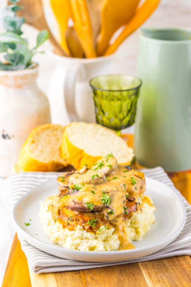 Plate of mashed potatoes topped with breaded meat and creamy gravy, garnished with herbs, served with slices of bread and a green glass in the background.
