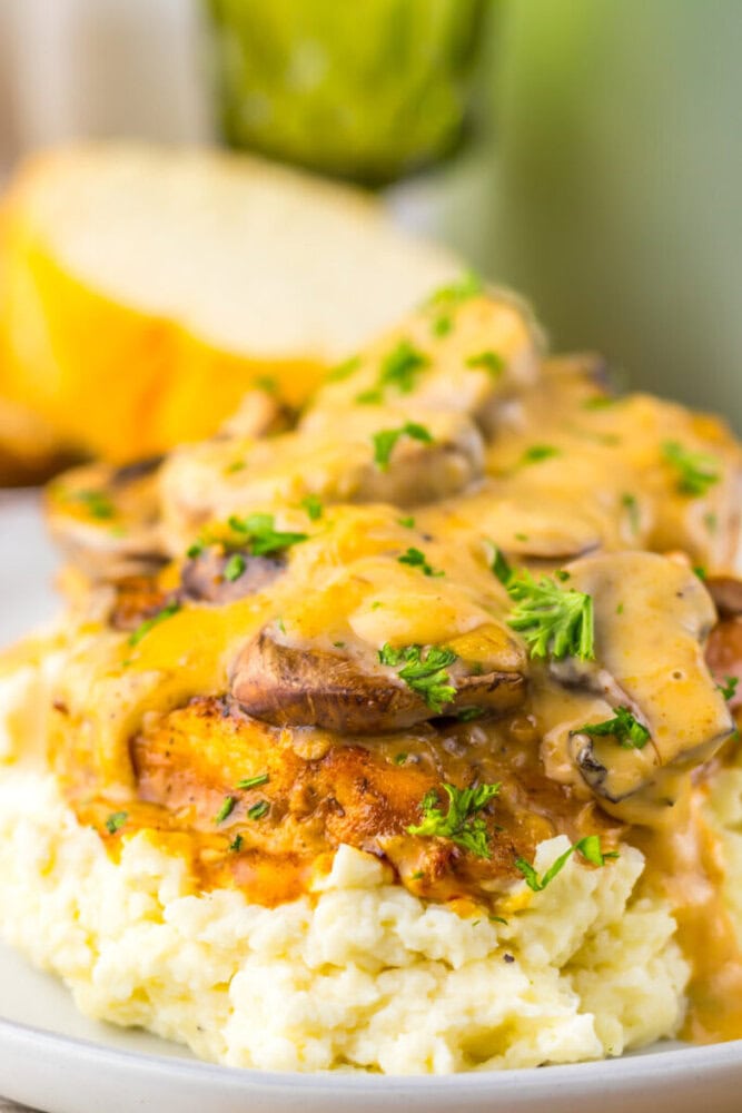 Grilled chicken breast topped with creamy mushroom sauce and chopped herbs, served over mashed potatoes, with a slice of bread in the background.
