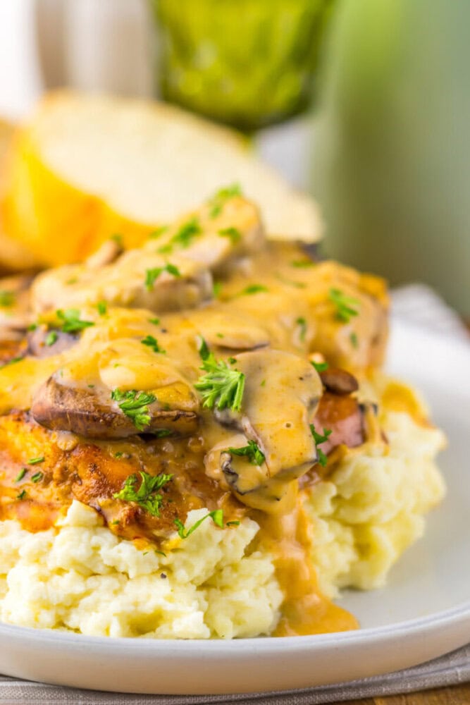 A plate of mashed potatoes topped with grilled chicken, sliced mushrooms, creamy sauce, and chopped herbs, with a slice of bread in the background.
