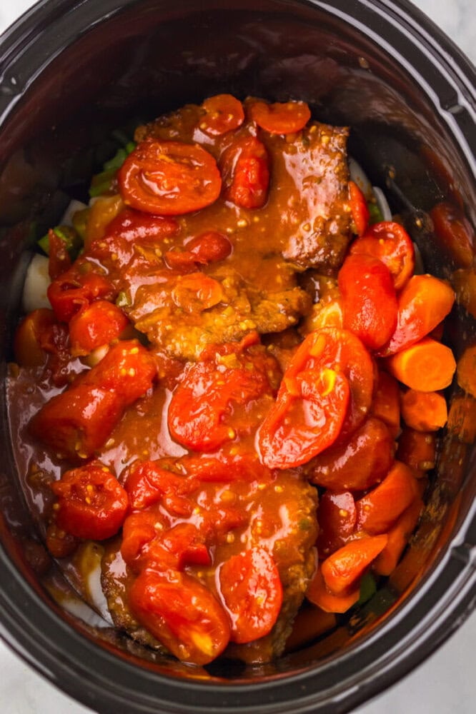 Sliced beef covered in tomato sauce with chopped tomatoes and carrots inside a slow cooker.