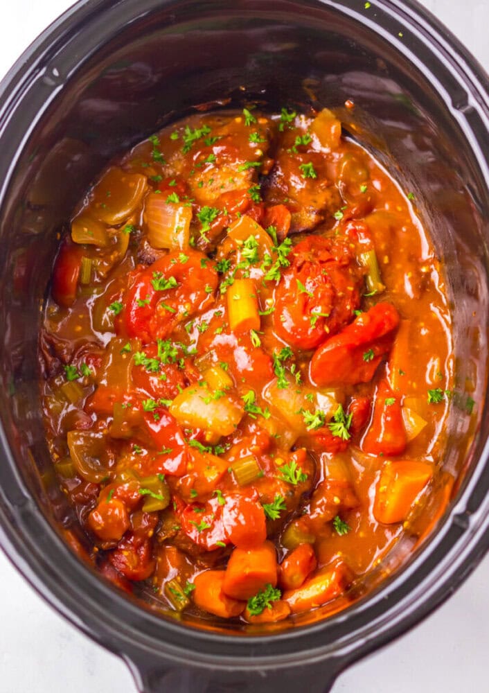 A close-up view of a slow cooker filled with beef stew, featuring chunks of beef, carrots, celery, and tomatoes in a thick sauce, garnished with chopped parsley.