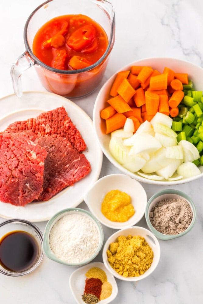 A variety of beef stew ingredients arranged on a white surface, including raw beef, canned tomatoes, chopped carrots, celery, onions, flour, seasonings, broth base, and soy sauce.