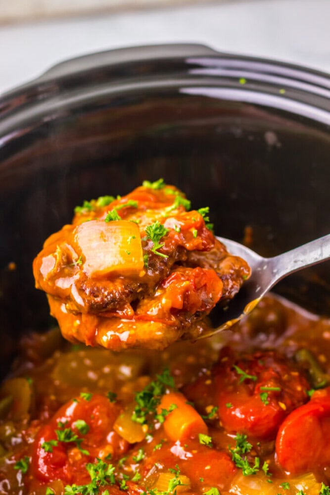 A spoonful of beef stew with vegetables and sauce is lifted from a slow cooker, garnished with chopped parsley.