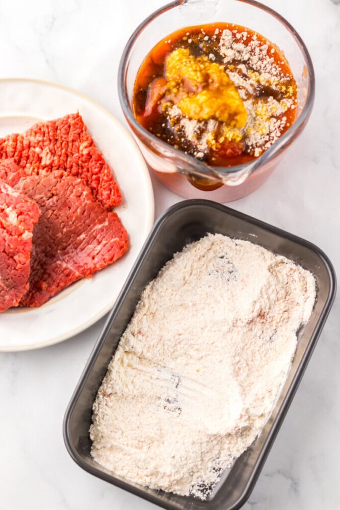 Raw beef slices on a plate, a container of seasoned flour, and a bowl of marinade ingredients on a white surface.