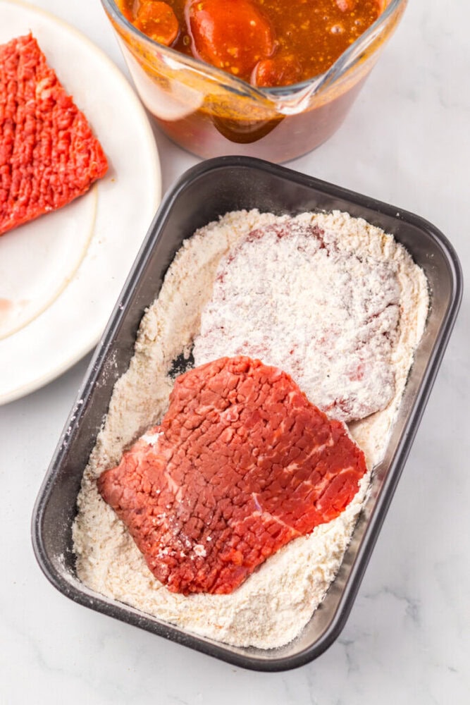 Two pieces of raw beef are being coated in flour in a black tray, with a plate of beef and a bowl of sauce nearby on a white countertop.